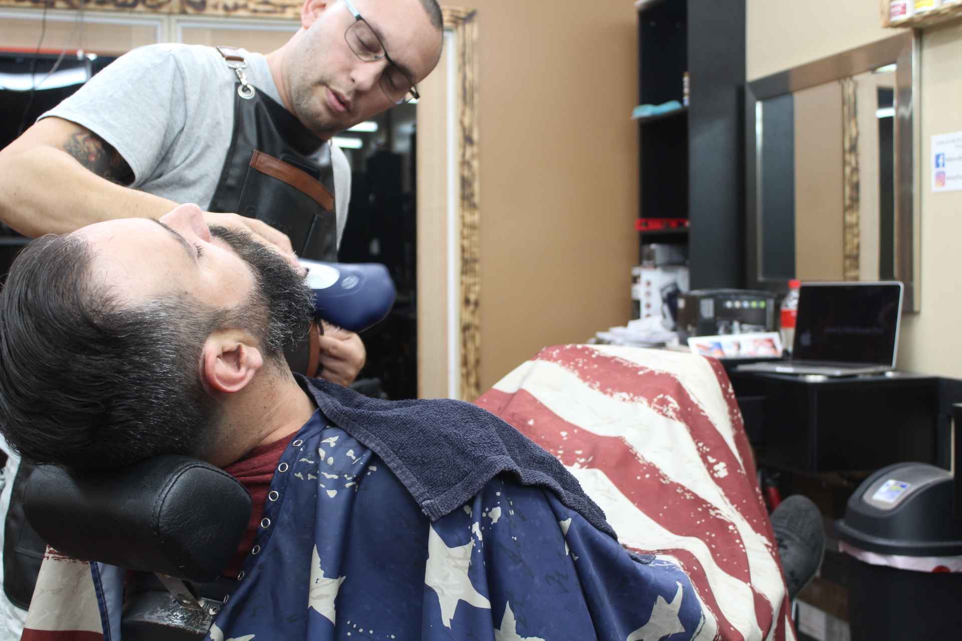 A barber grooming a reclined client’s beard in the studio chair