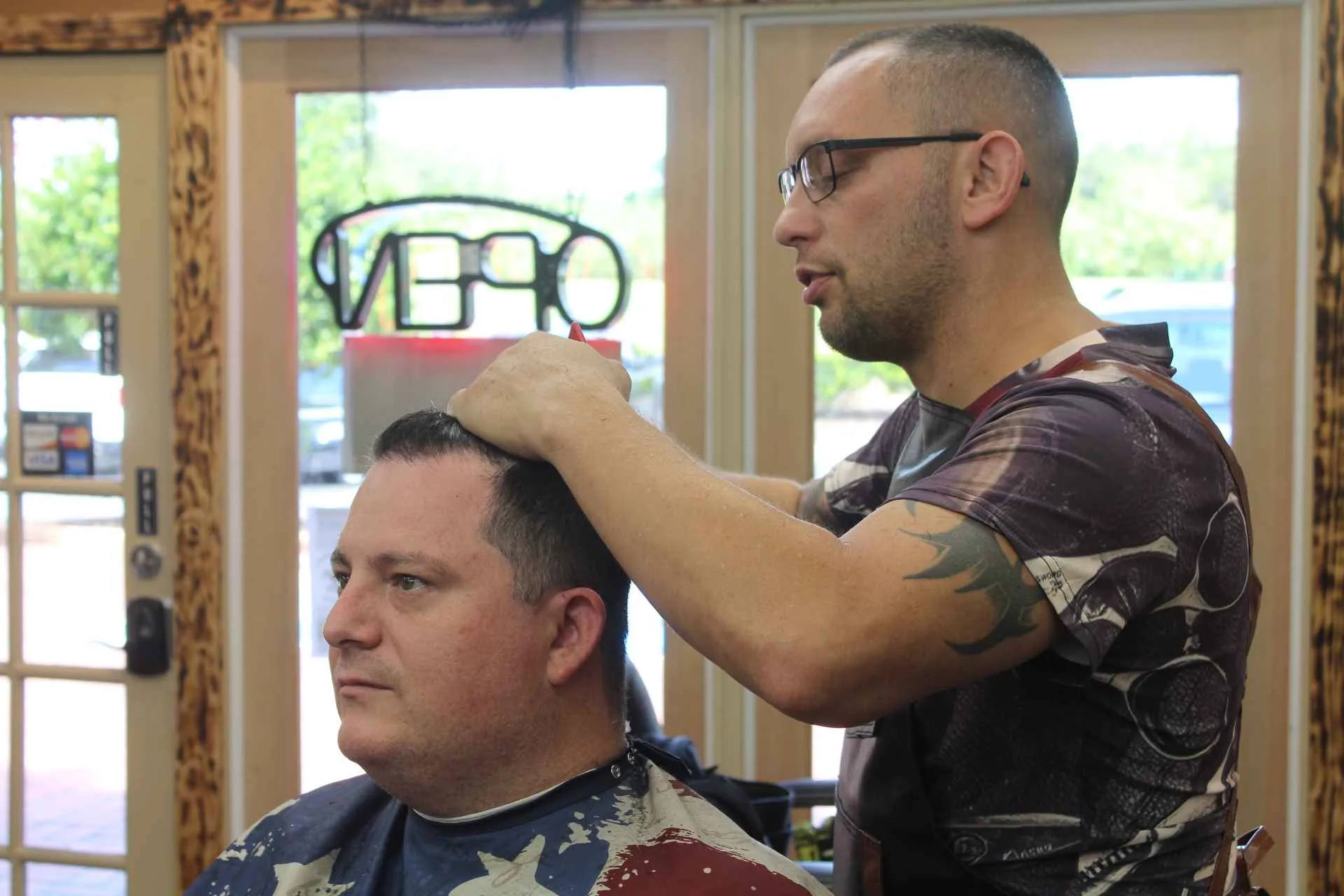 A barber shaping a seated client’s hair beside the studio’s front windows and open sign